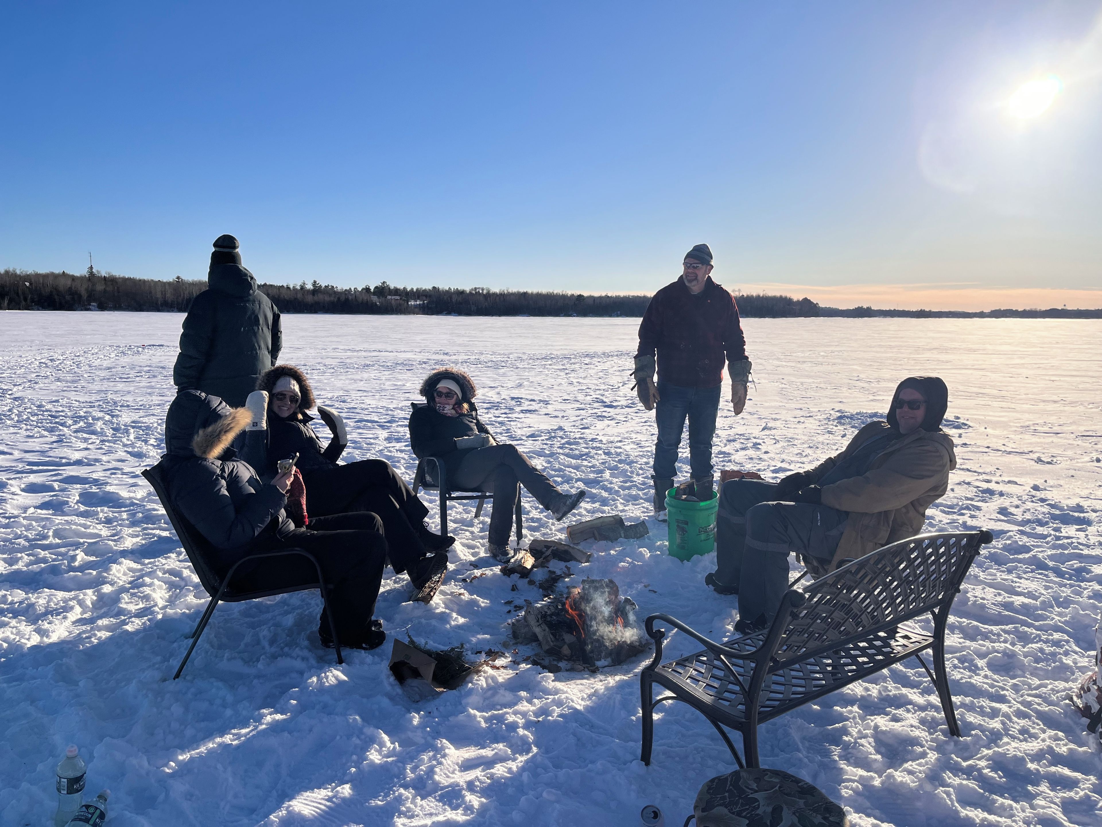 A group of people sitting and standing on a snow-covered field with a bench and a fire pit.