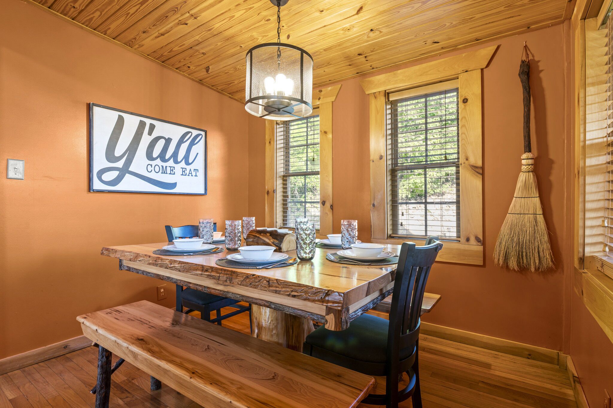 Un coin repas rustique avec une table en bois dressée pour quatre personnes, un luminaire suspendu et un balai accroché au mur.