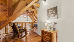 Et hyggeligt sommerhusinteriør med træbjælker, en gyngestol, et træbord med en lampe, og et skilt med teksten 'Cabin Sweet Cabin'.