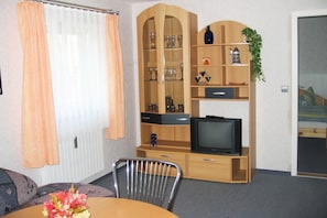 Ein Wohnzimmer mit Holzschrank, Fernseher, Esstisch mit Stühlen und einem fenster mit Vorhängen.