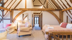 Una habitación acogedora con techo alto y vigas de madera a la vista, con dos camas con ropa de cama rosa, un sofá blanco y una mesita con una lámpara.