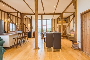 A spacious room with a wooden floor, a dining area with a long table and chairs, and a kitchen area with a counter and stools. - The Canterbury Barn by Bloom Stays - large barn sleeping up to 20. Ideal for hen parties (Canterbury,)