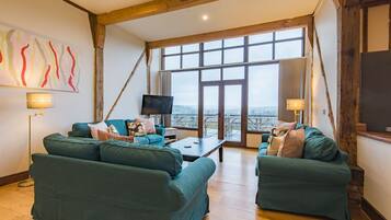 A spacious living room with two green sofas, a coffee table, and a TV. Large windows offer a view of the outdoors.