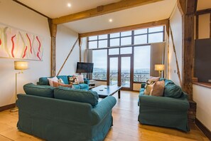 A spacious living room with two green sofas, a coffee table, and a TV. Large windows offer a view of the outdoors.