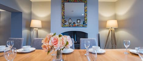 Ein Esszimmer mit Holztisch, gedeckt für sechs Personen, flankiert von zwei Lampen, einem Spiegel mit farbenfrohem Rahmen und einer Blumenvase.