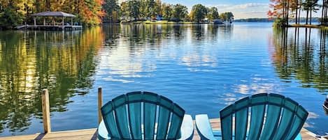 A wooden dock with two chairs facing a calm lake, surrounded by trees and a clear sky.