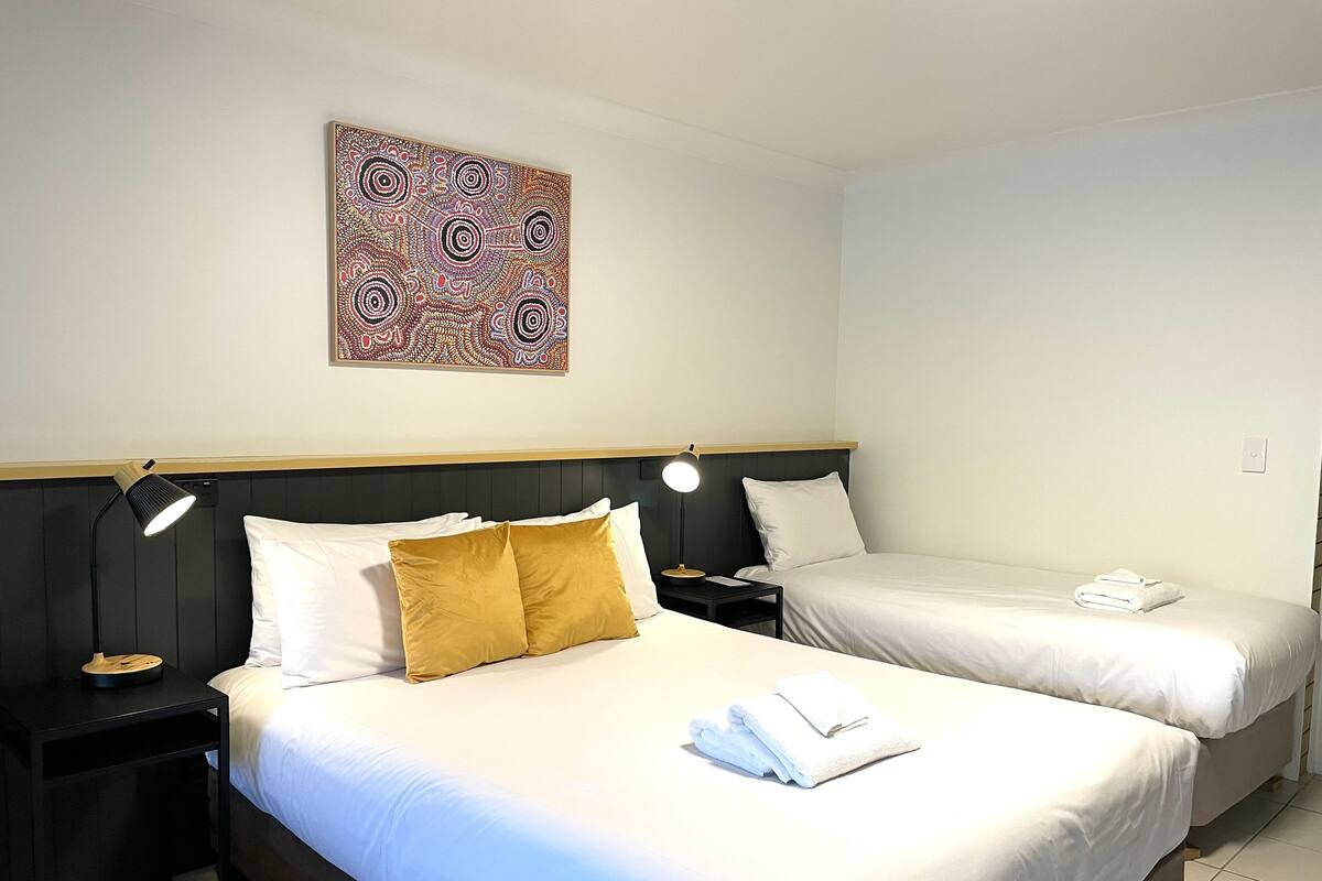 Habitación de hotel con dos camas, una mesita de noche con lámpara, una obra de arte en la pared y una toalla doblada sobre la cama.