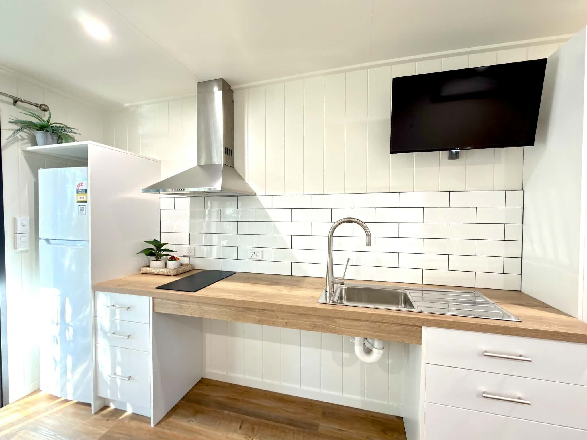 Una cocina moderna con gabinetes blancos, extractor de acero inoxidable, pileta integrada y un televisor de pantalla plana sobre la encimera.
