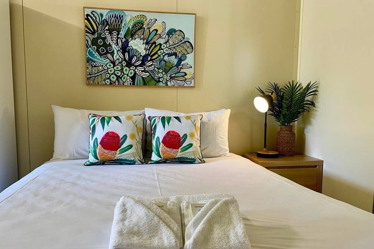 Una cama bien hecha con dos almohadas decorativas, una obra de arte en la pared, una planta en la mesita de noche y una lámpara.