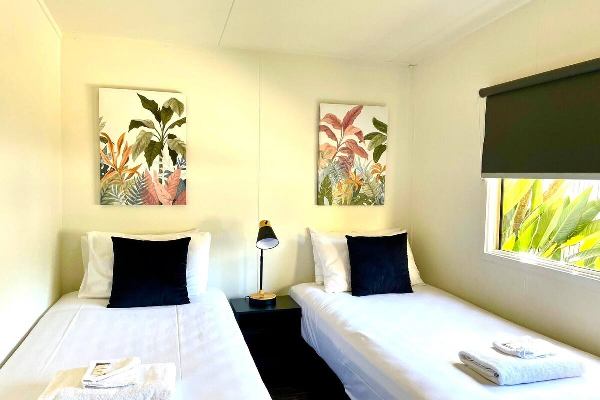 Habitación de hotel con dos camas individuales, cada una con una almohada y una toalla doblada. Hay una mesita de noche con una lámpara entre las camas.