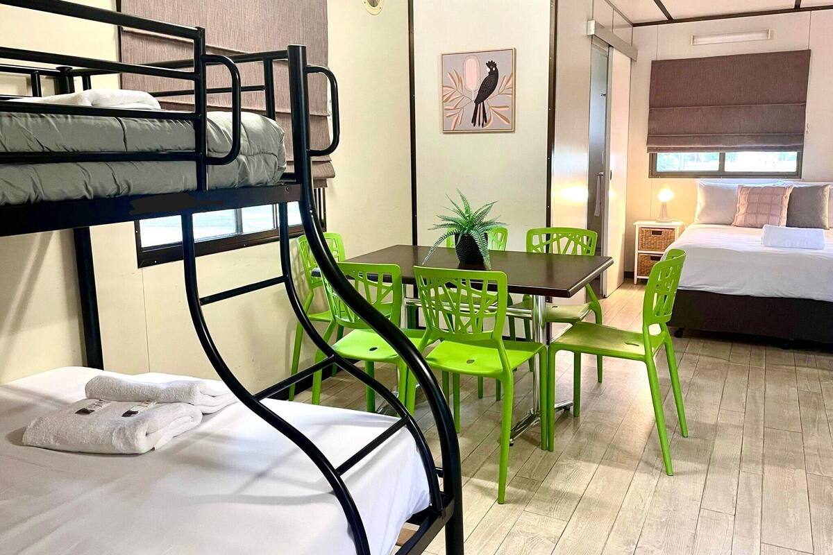 Una cama litera con ropa blanca, una mesita con una planta, sillas verdes, y una cama con ropa blanca en una habitación con piso de madera y una ventana.