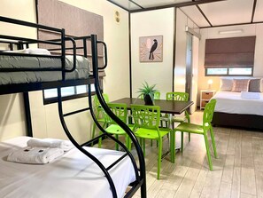 A bunk bed with white bedding, a small table with a plant, green chairs, and a bed with white bedding in a room with wooden flooring and a window.