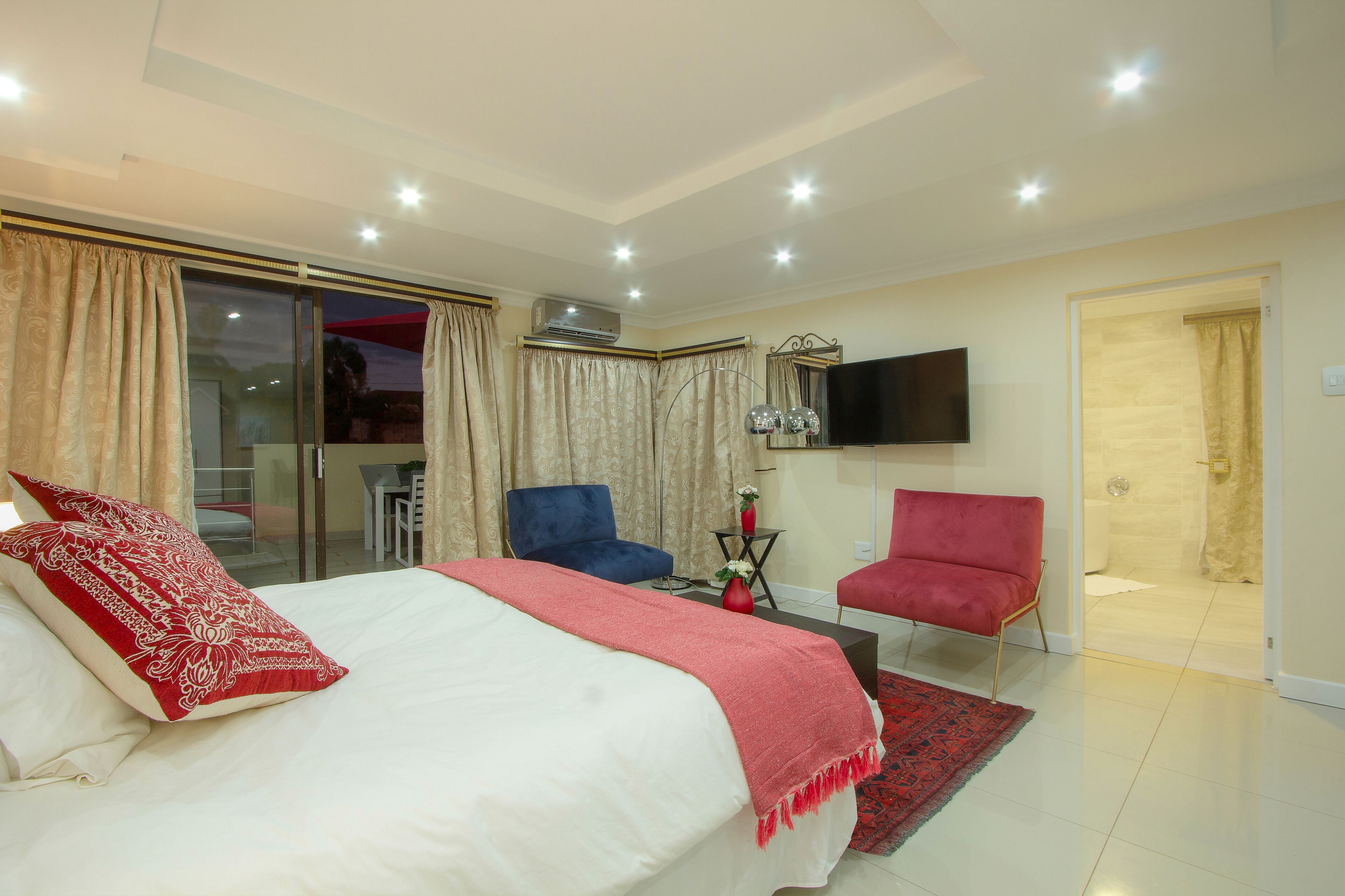 A hotel room with a large bed, a red and white patterned pillow, a blue chair, a red chair, a small table with a vase, and a TV mounted on the wall.