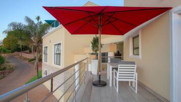 A balcony with a red umbrella, white furniture, and a view of a road and greenery.