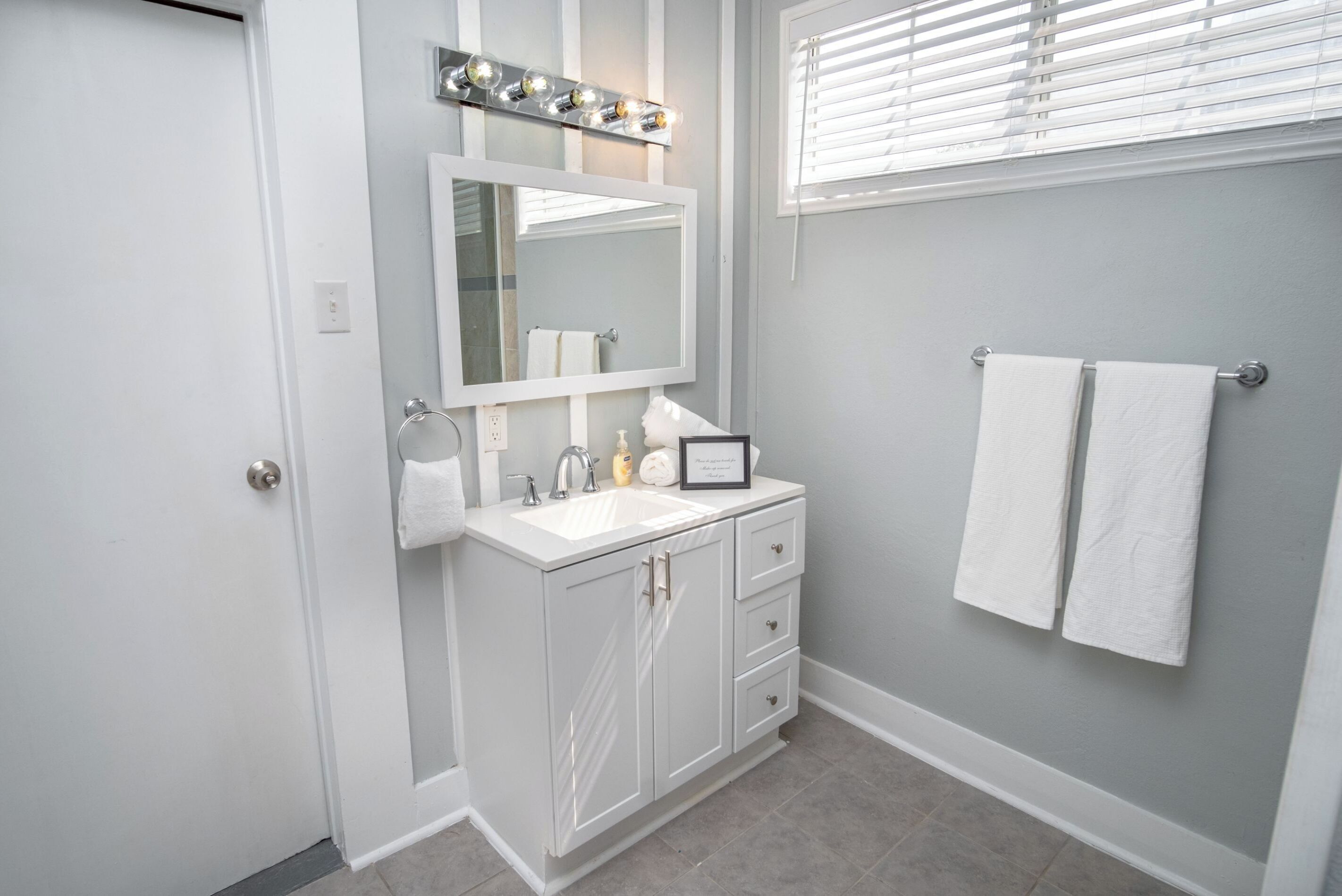 A bathroom with a white vanity, a large mirror with lights, and two towels on the wall.