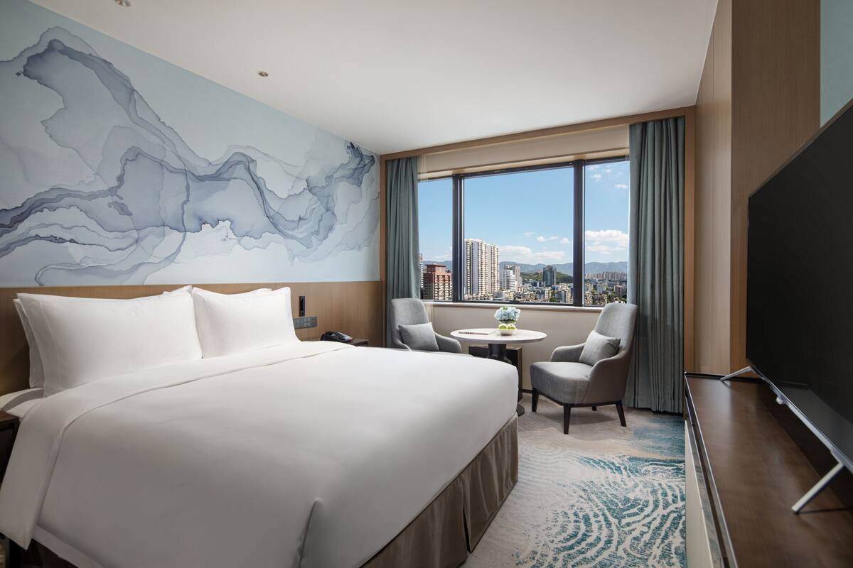 Ein Hotelzimmer mit einem großen Bett, einem Schreibtisch, einem Sessel, einem Fernseher und Blick auf die Stadt.