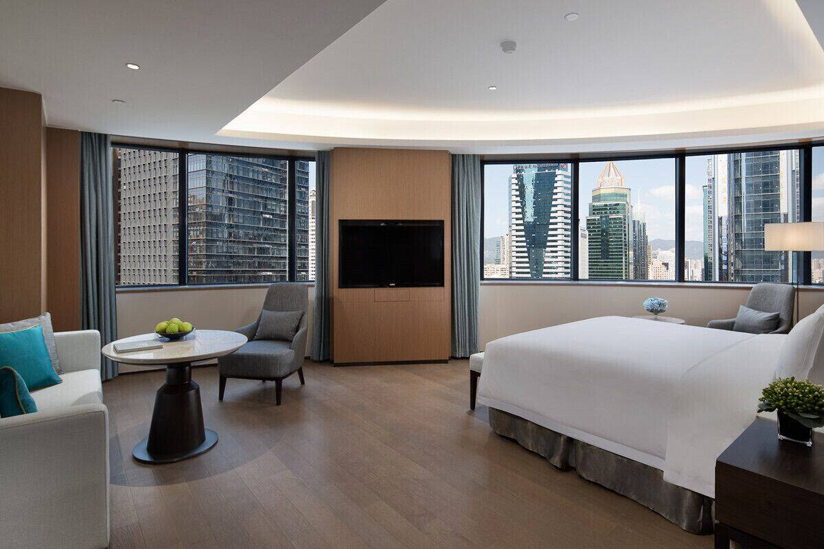 Ein modernes Hotelzimmer mit einem großen Bett, einem Fernseher, einer Sitzecke mit Tisch und Blick auf die Stadt durch große Fenster.