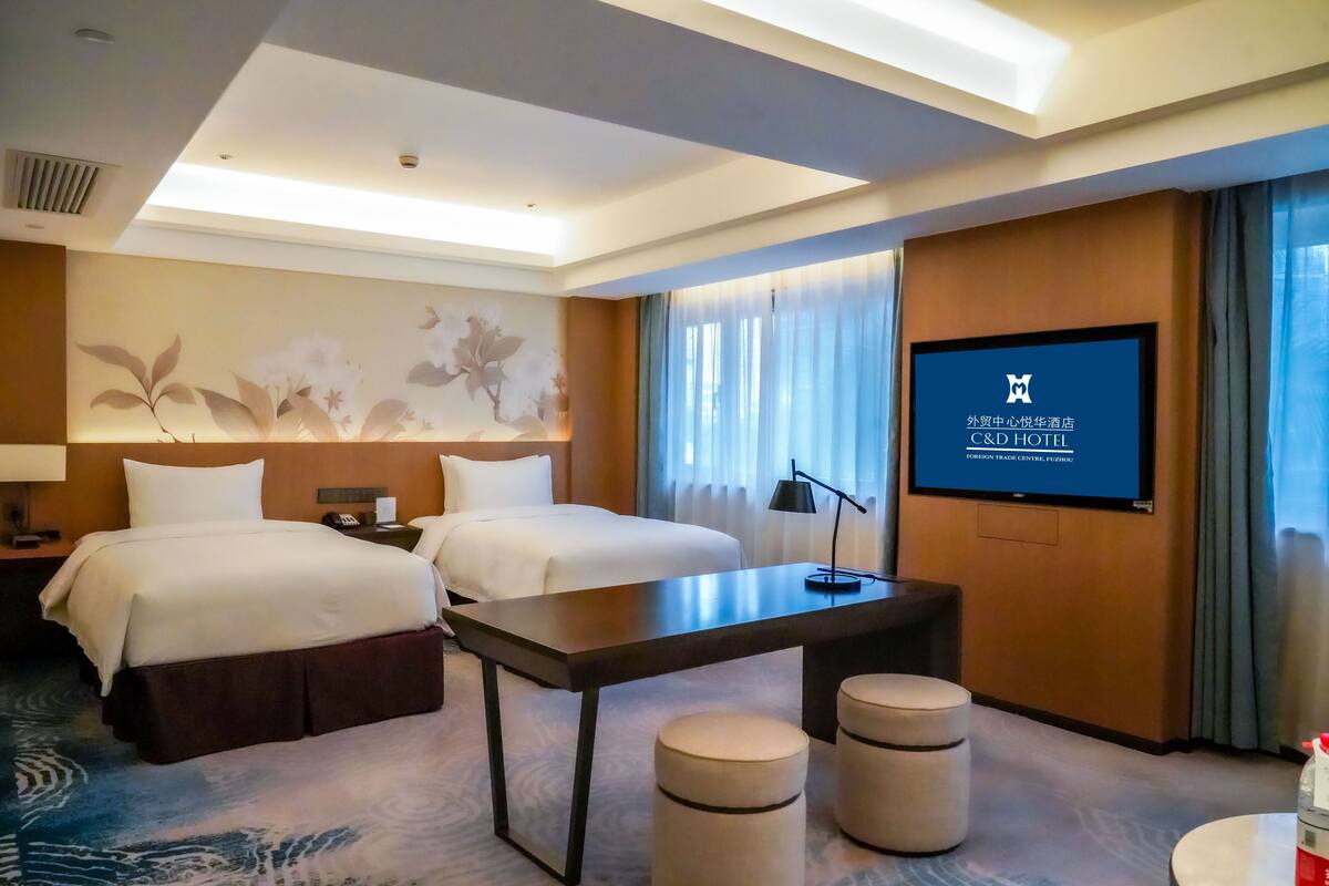Ein Hotelzimmer mit zwei Betten, einem Schreibtisch, einem Fernseher und einem großen Fenster mit Vorhängen.