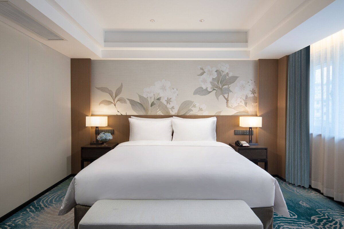 Une chambre d’hôtel avec un grand lit, deux tables de chevet avec des lampes, une œuvre d’art accrochée au mur et une grande fenêtre avec des rideaux.
