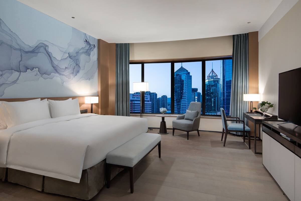 Ein Hotelzimmer mit einem großen Bett, einem Schreibtisch mit Stuhl, einem Fernseher und Blick auf eine Stadtkulisse durch große Fenster.
