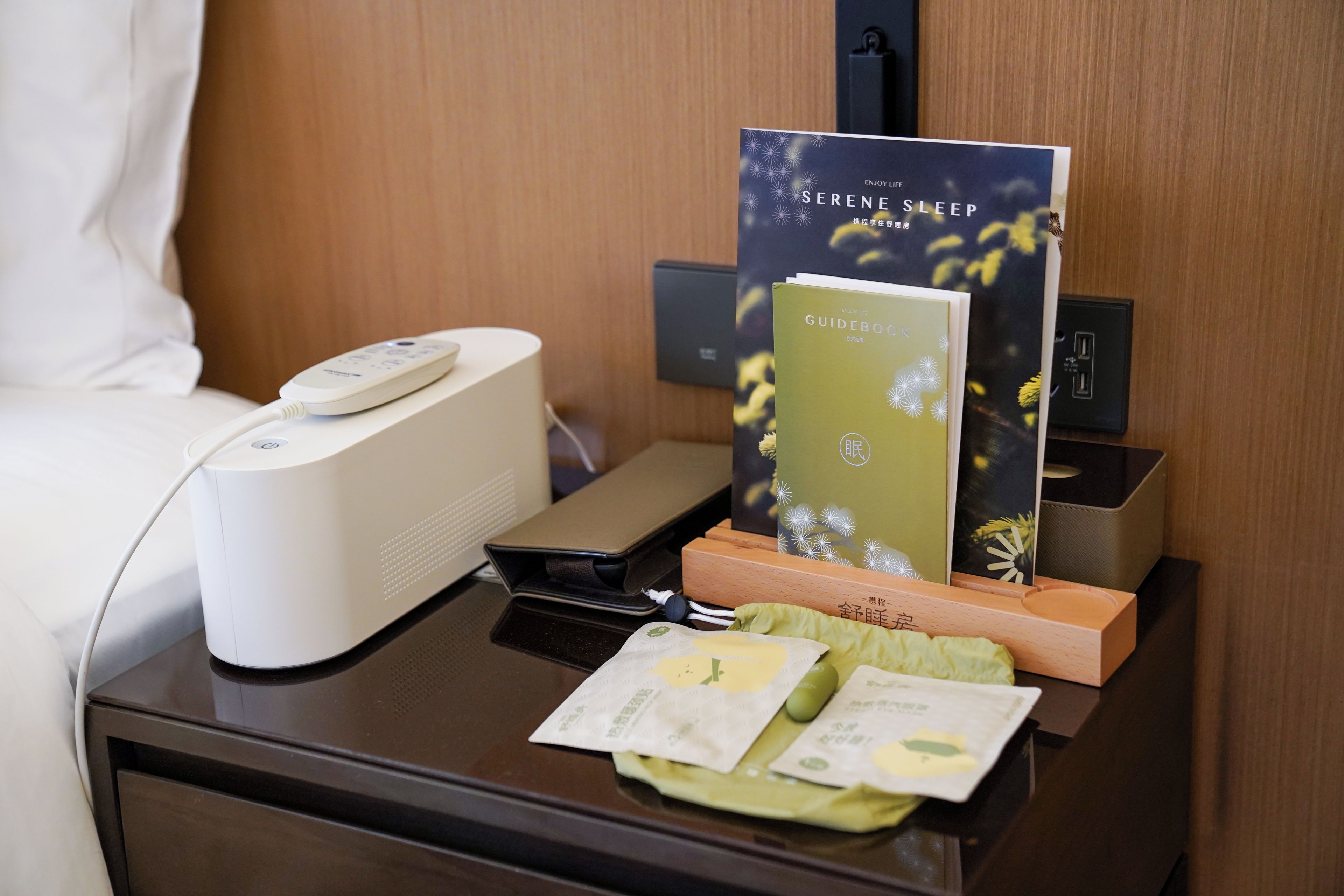 A hotel room with a bedside table featuring a remote control, a book titled 'Serene Sleep,' a tissue box, and a pair of earphones.