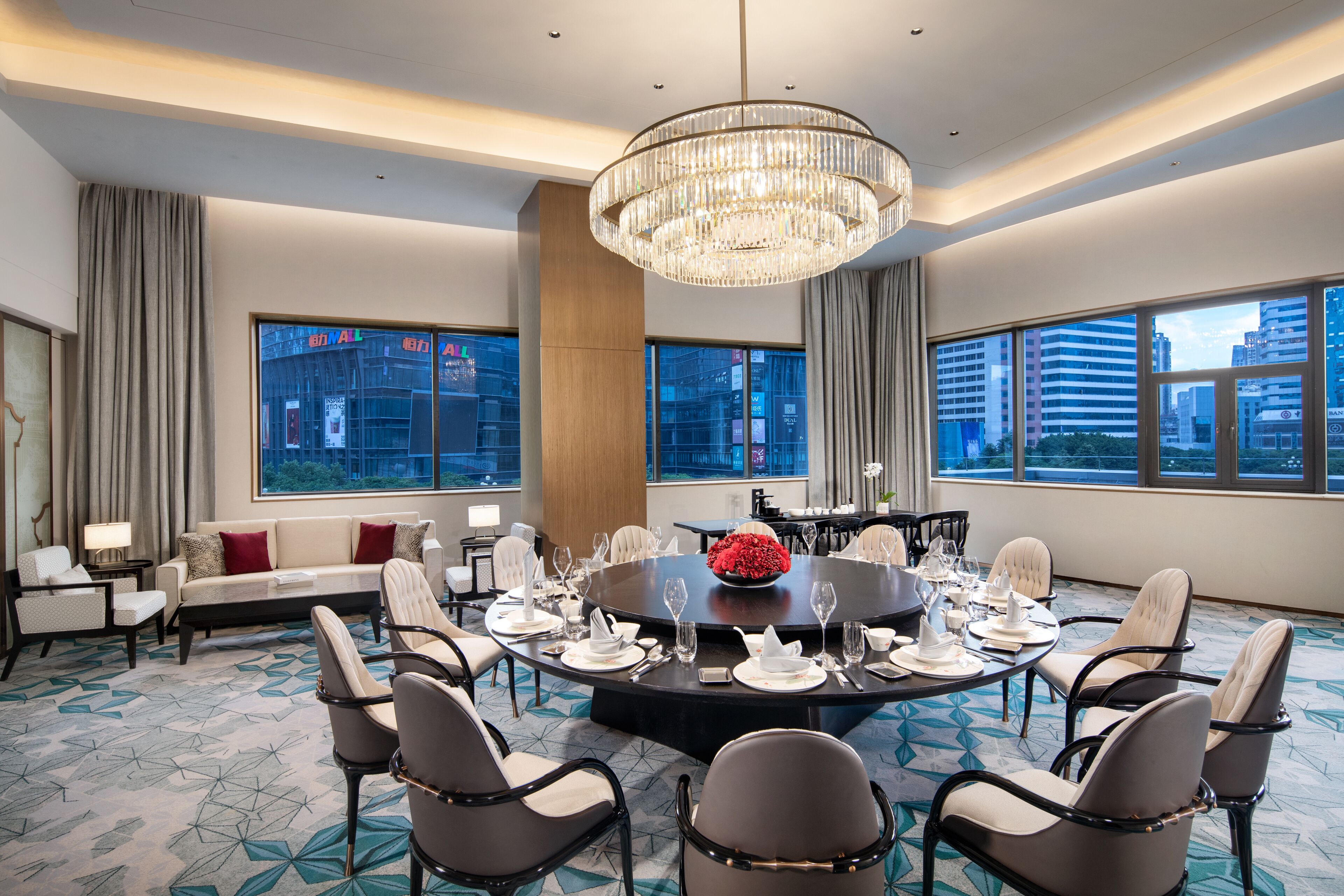 A modern hotel conference room with a round table set for dining, large windows with city views, and a chandelier.