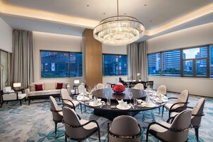 A modern hotel conference room with a round table set for dining, large windows with city views, and a chandelier.