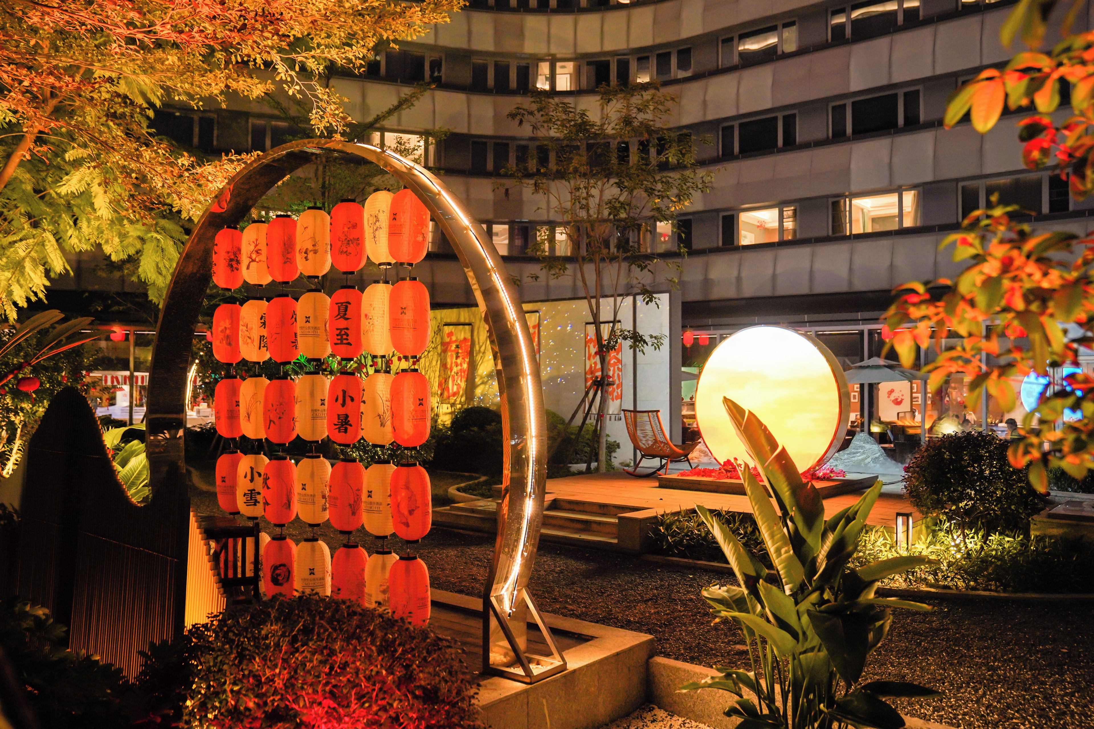 Une cour intérieure dotée d’une grande structure en arc sur laquelle sont suspendues des lanternes, entourée de plantes et avec un bâtiment en arrière-plan.