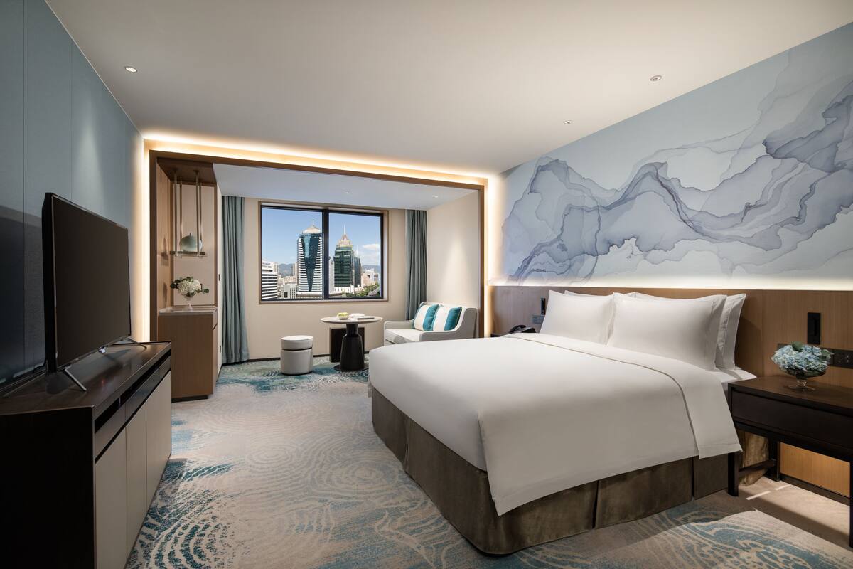 Ein modernes Hotelzimmer mit einem großen Bett, einem Fernseher, einem Schreibtisch und Blick auf die Stadt.