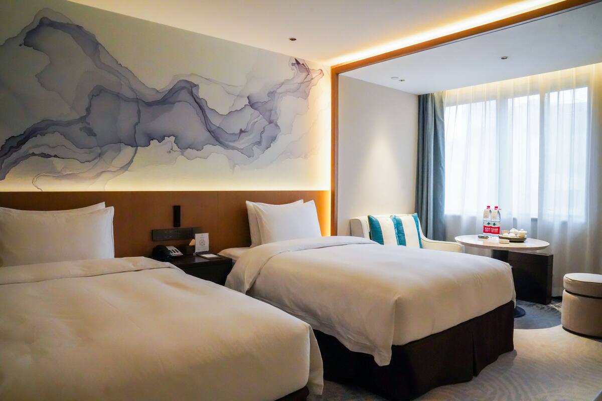 Ein Hotelzimmer mit zwei Betten, einem großen abstrakten Gemälde an der Wand, einem runden Tisch mit Flasche und Gläsern sowie Blick ins Freie durch das Fenster.