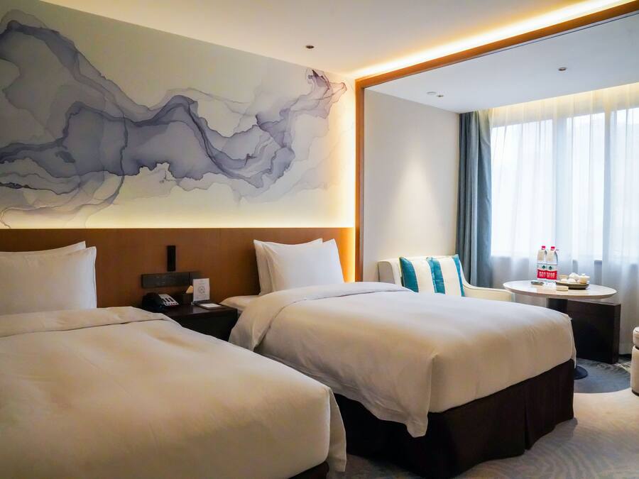Habitación de hotel con dos camas, un cuadro abstracto grande en la pared, una mesa redonda con una botella y vasos, y vista al exterior a través de la ventana.