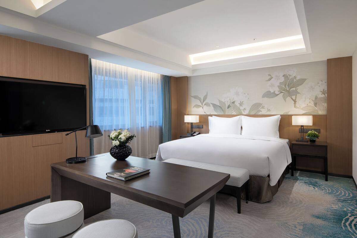 Ein Hotelzimmer mit einem großen Bett, einem Flachbildfernseher, einem Schreibtisch mit Lampe und einer Wanddekoration.