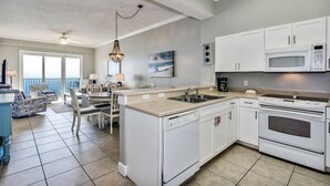Una cocina moderna con gabinetes blancos, lavavajillas integrado, microondas y horno. Hay una zona de comedor con mesa y sillas, y un ventanal con vista al océano.