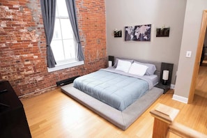 Een slaapkamer met een bakstenen muur, een groot bed, een raam met gordijnen en een wereldkaart aan de muur.