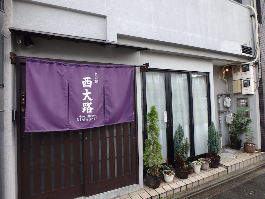 A building with a purple banner reading "Guest House Nishioji" and a brown gate. There are potted plants on the sidewalk.