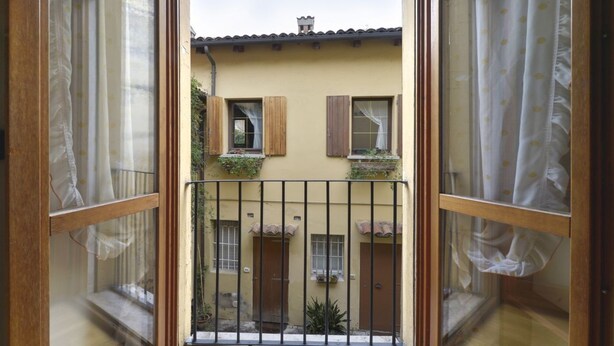 Blick durch ein Fenster mit Holzrahmen und Vorhängen auf ein Gebäude mit gelber Fassade, Holzhörnern und einem Balkon mit Pflanzen.
