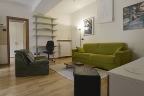 A modern living room with a green sofa, a green armchair, a black ottoman, and a white desk. There is a radiator, a floor lamp, and a painting on the wall. - UR-NEST Sant' Apollonia (Bologna)