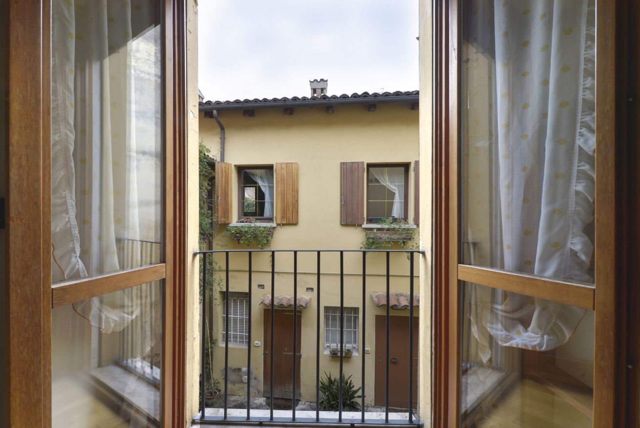 A view through a window with a wooden frame and curtains, looking out at a building with a yellow facade, wooden shutters, and a balcony with plants.