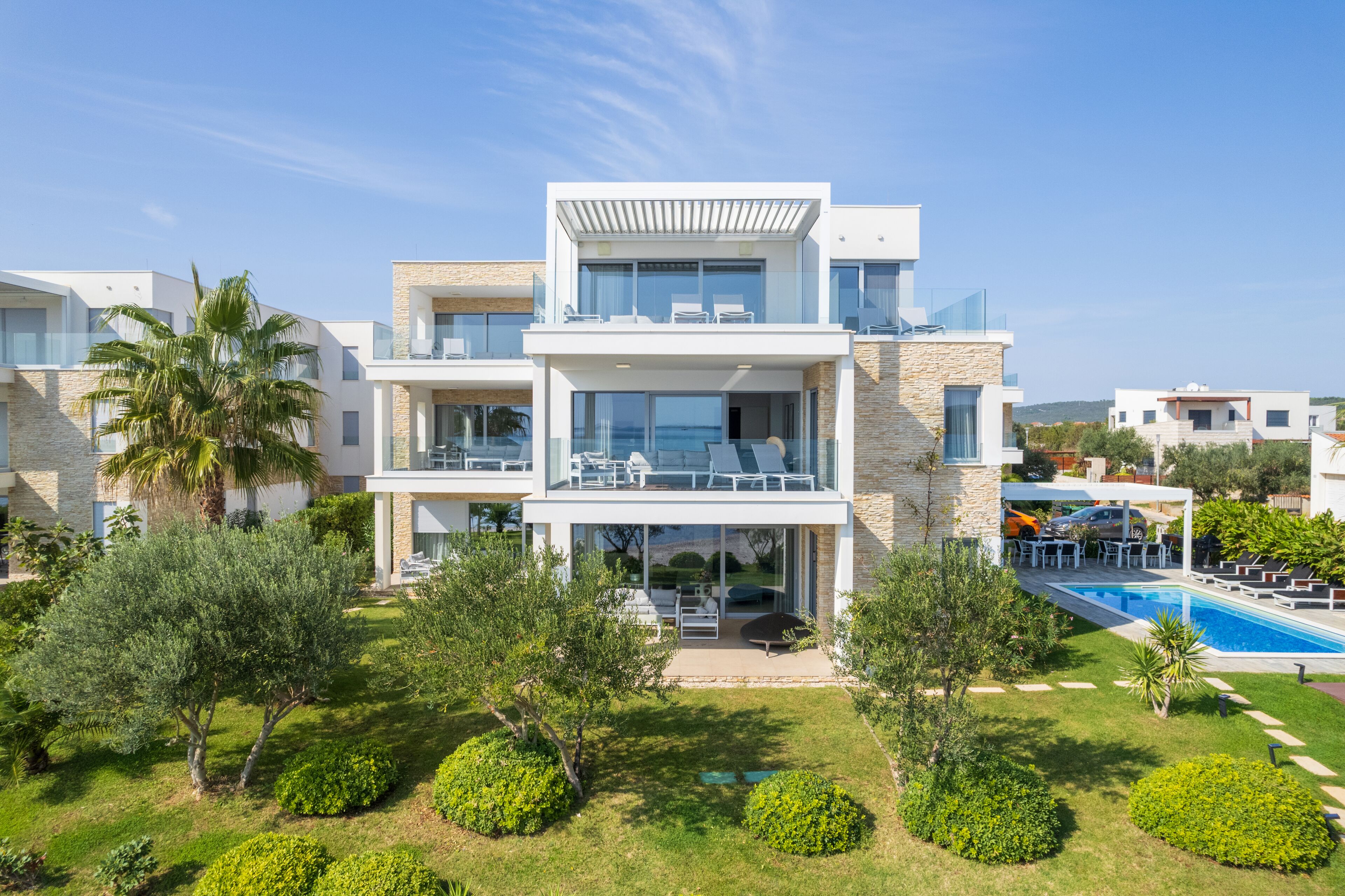 A modern multi-story house with a pool, surrounded by lush greenery and palm trees.