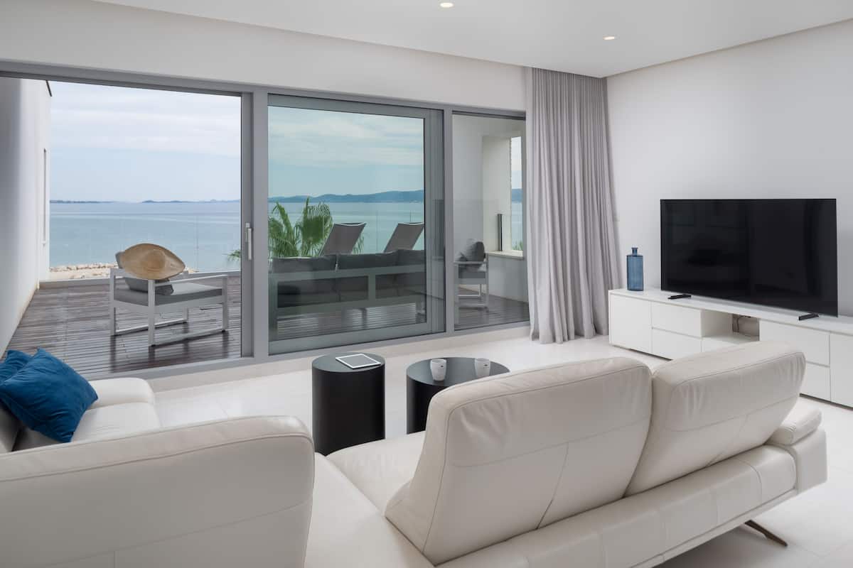 Una sala de estar moderna con televisor grande, sofá blanco y balcón con vista al mar.
