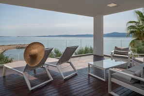 Una terraza de madera con mobiliario blanco, incluyendo dos sillones reclinables y un sofá, con vistas a una masa de agua tranquila y montañas distantes.