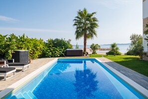 Ein rechteckiger Swimmingpool mit glasklar blauem Wasser, umgeben von einer Holzterrasse. Auf der Terrasse stehen Liegestühle und ein kleines Holzgebäude. Im Hintergrund sind eine Palme und verschiedene Sträucher zu sehen.