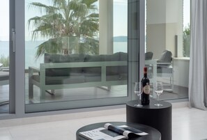 Ein modernes Zimmer mit einem großen Panoramafenster, das Blick auf einen Strand und Palmen bietet. Im Vordergrund steht ein schwarzer runder Tisch mit einer Weinflasche und zwei Gläsern.