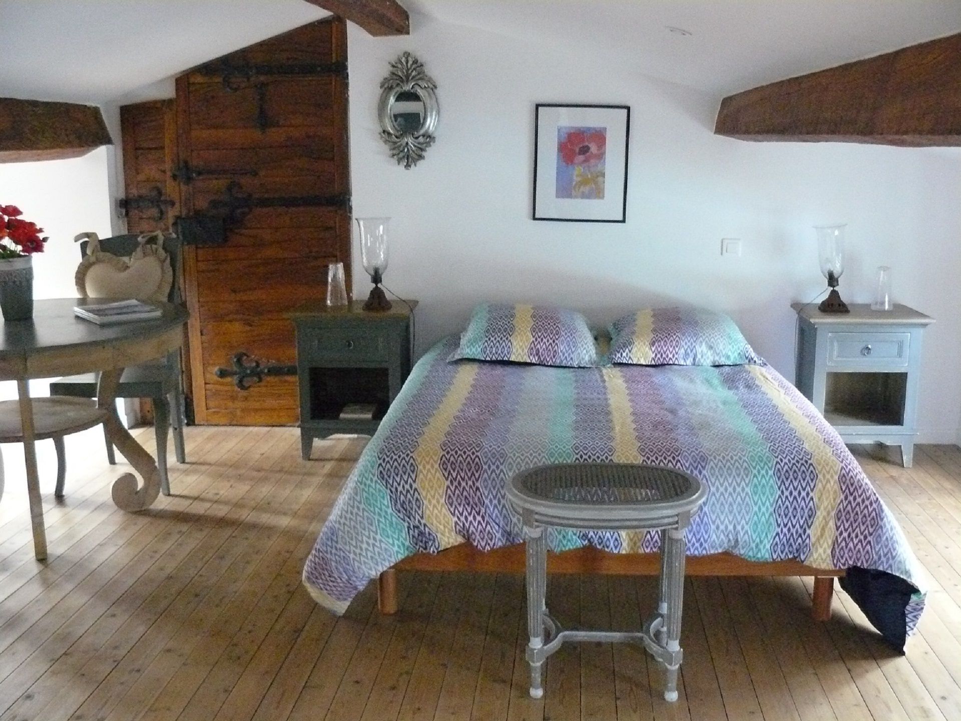 Une chambre avec un plancher en bois, un lit avec une literie à motifs, une petite table, une chaise, une commode et un miroir fixé au mur.