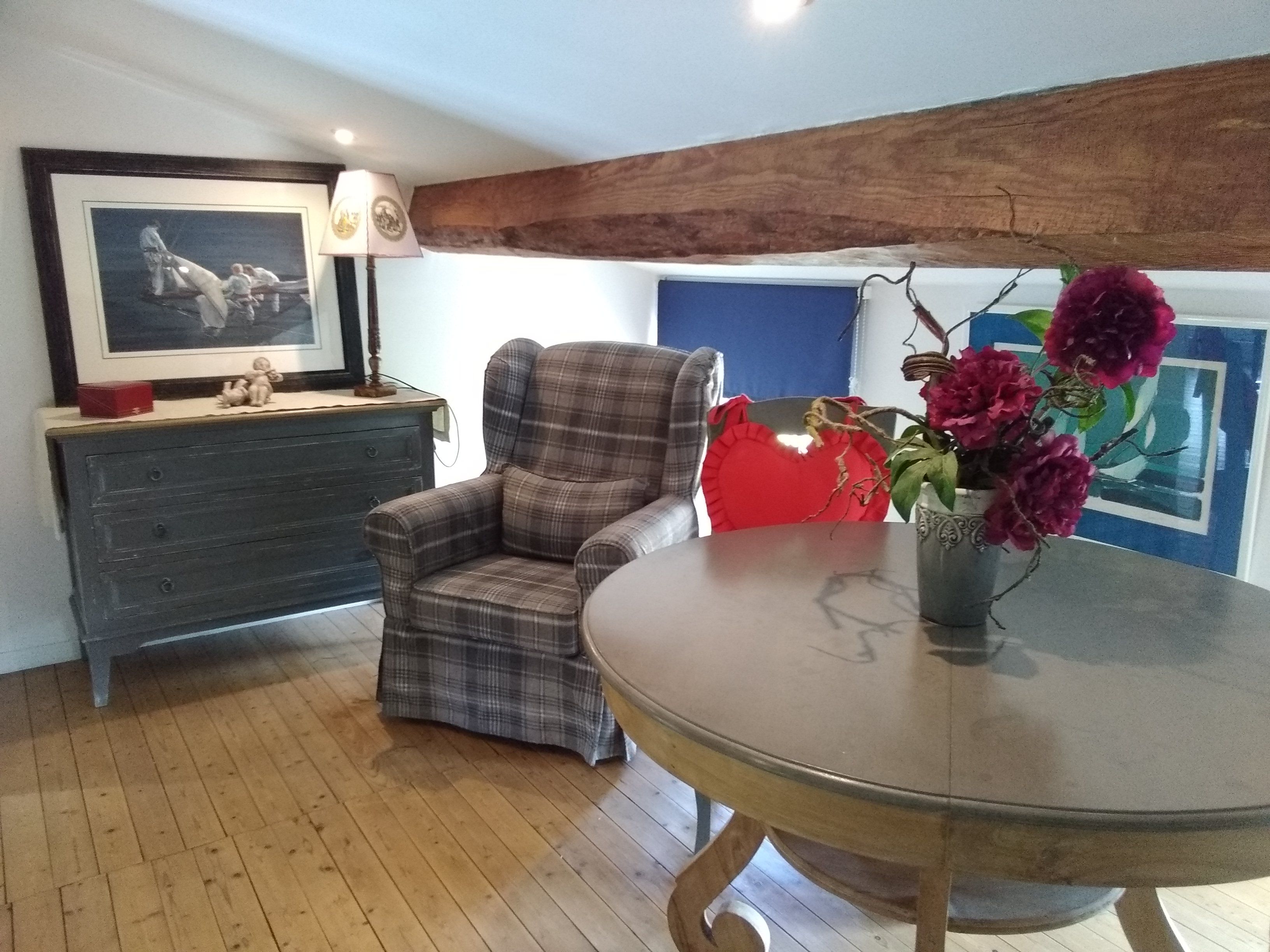 A room with a plaid armchair, a round table with a vase of flowers, a dresser with a framed picture, and a blue wall with a painting.