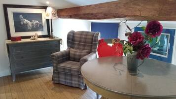 A room with a plaid armchair, a round table with a vase of flowers, a dresser with a framed picture, and a blue wall with a painting.