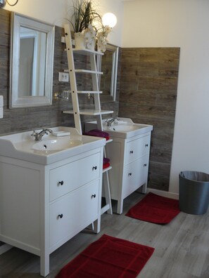 A bathroom with two sinks, a mirror, a shelf with plants, and a red rug.