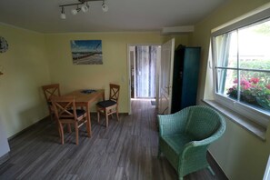 Ein Essbereich mit Tisch und Stühlen, ein grüner Korbstuhl, ein Bild an der Wand und ein Fenster mit Pflanzen.