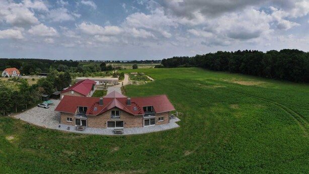 Une grande maison de plain-pied, avec un toit rouge, entourée d’un champ verdoyant et d’une forêt.