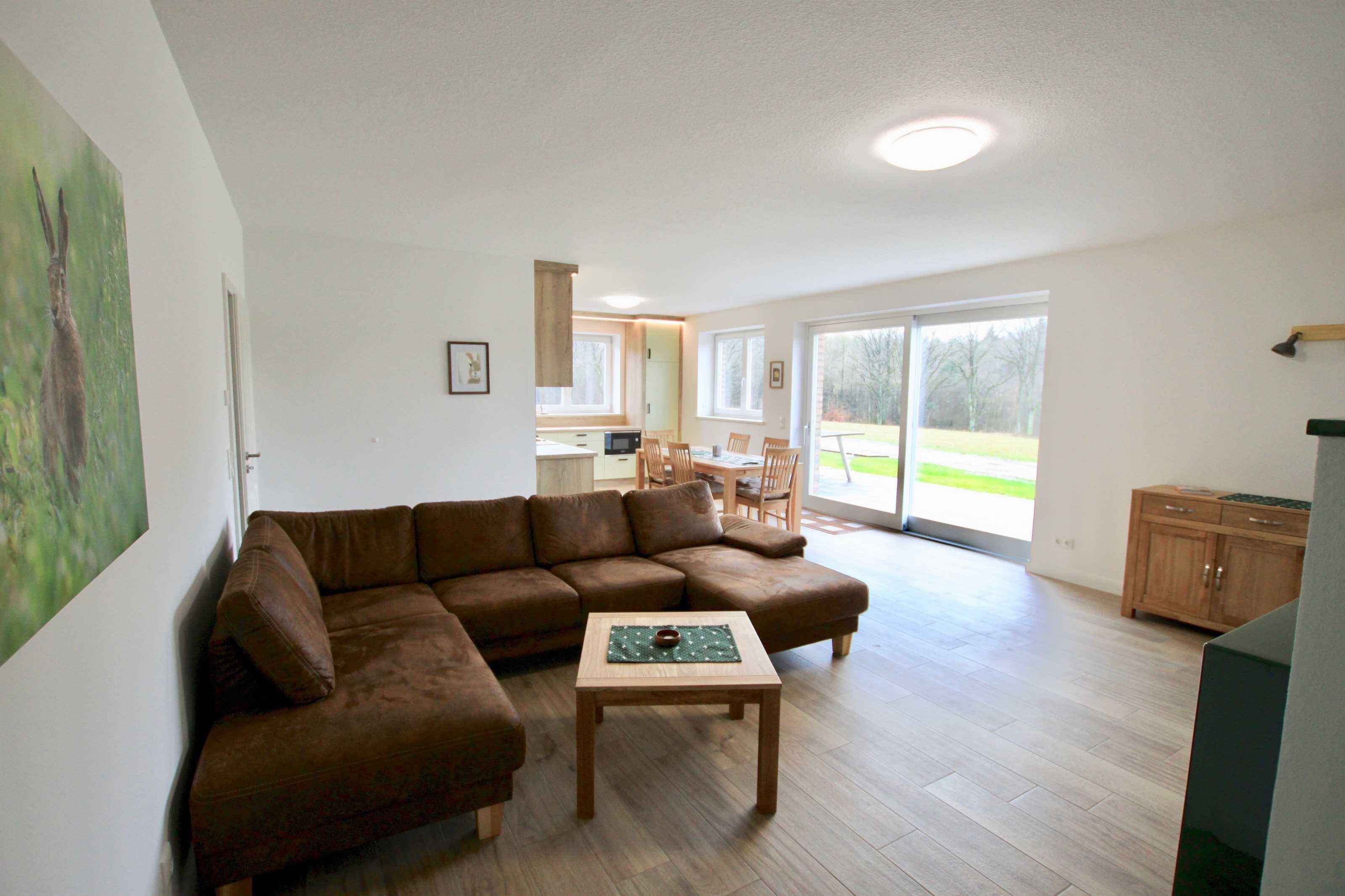 A living room with a brown sectional sofa, a wooden coffee table, and a kitchen area in the background.